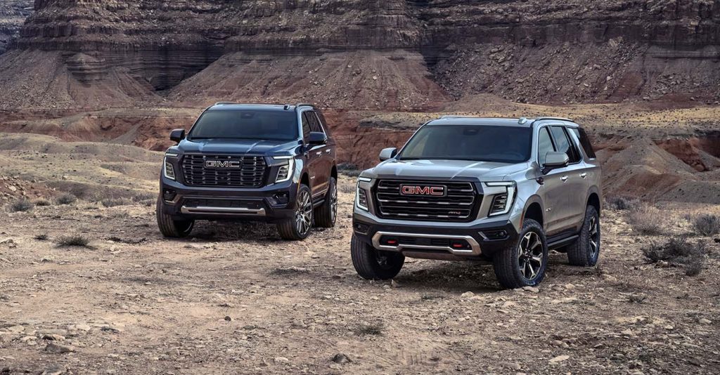 Two 2025 GMC Yukon SUVs parked on rugged desert terrain with rocky cliffs in the background, showcasing their bold front grilles, modern LED headlights, and off-road-ready design.