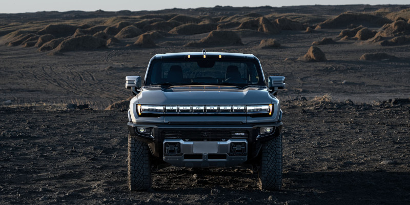 Front view of the 2025 GMC HUMMER EV Pickup on rugged terrain, highlighting its bold LED lighting and off-road stance.